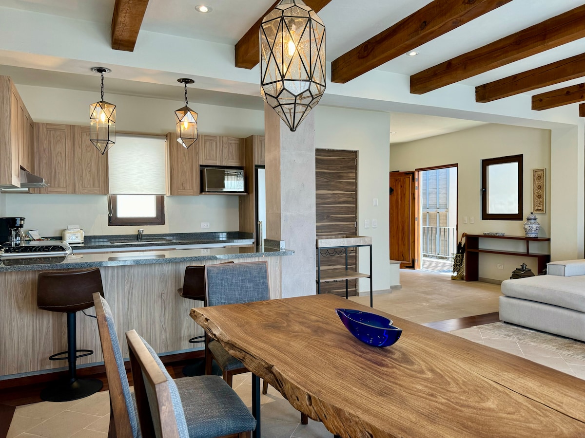 An inviting dining area features a rustic wooden table accompanied by stylish chairs. Modern pendant lights hang above, illuminating the space. The open kitchen is visible in the background, showcasing sleek cabinetry and countertop appliances, while natural light streams through the nearby doorway.