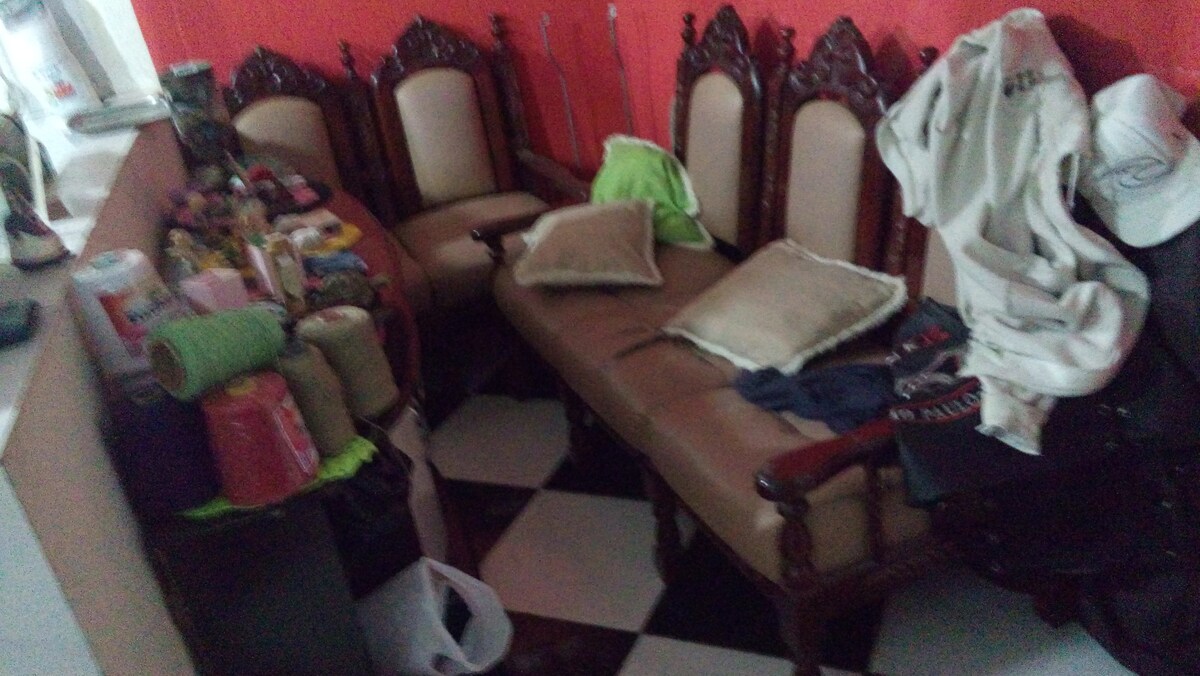 A seating area is furnished with a brown sofa and several ornate wooden chairs. Various items are placed around the furniture, including pillows, clothing, and colorful rolls, creating a lived-in atmosphere. The floor features a contrasting black and white checkerboard pattern.