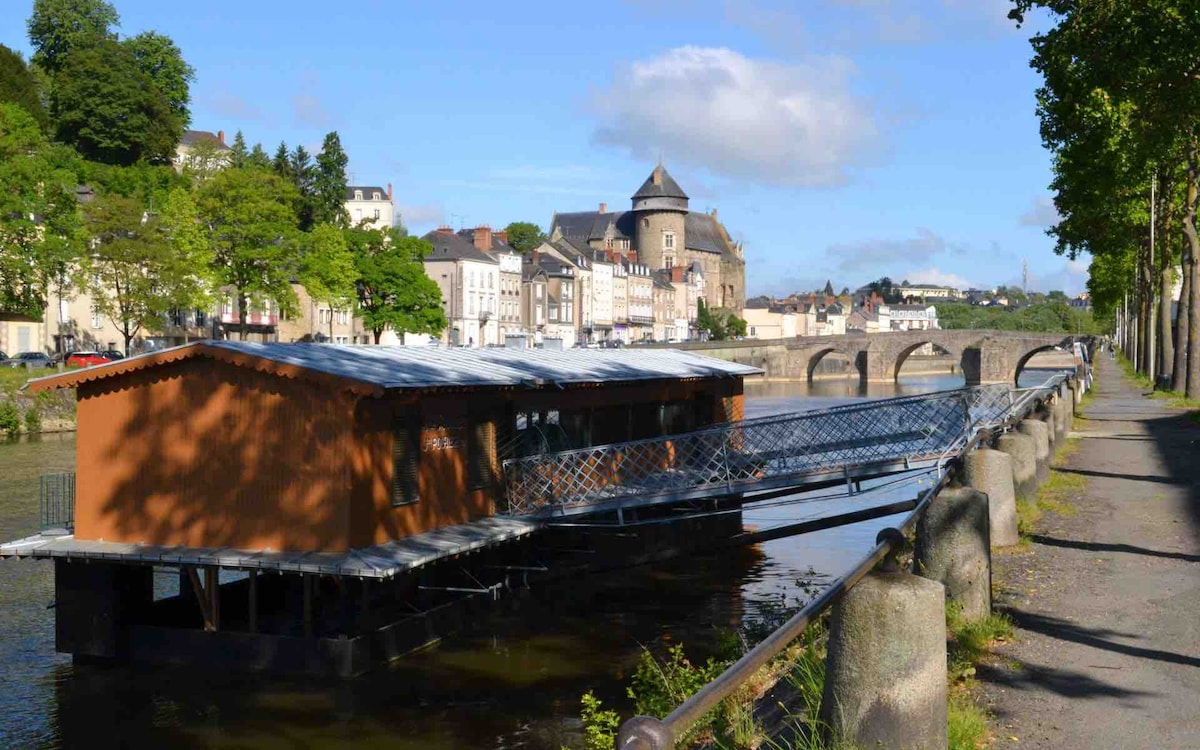 A scenic view features a floating structure positioned on a calm river alongside a tree-lined pathway. Historic buildings rise in the background, with a castle-like structure visible. The scene is framed by vibrant greenery and a clear blue sky, enhancing the natural setting.