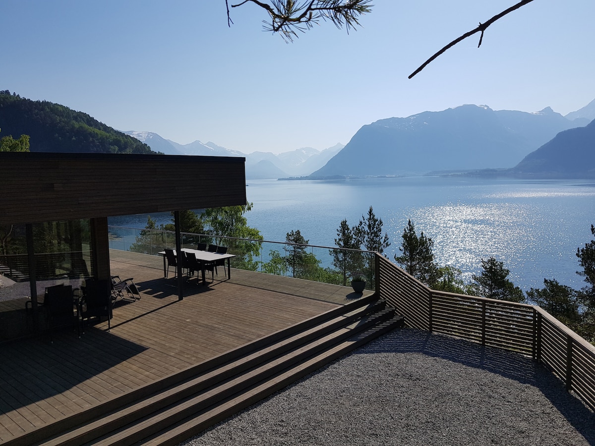 A spacious outdoor deck offers stunning views of a calm lake surrounded by mountains. The deck features a dining table and lounge chairs, while the gravel area adds texture. Sunlight reflects on the water, enhancing the serene atmosphere of the location.