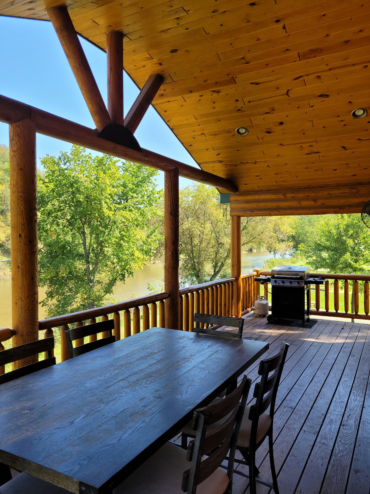 An outdoor deck features a wooden dining table surrounded by six chairs, ideal for family meals. A charcoal grill sits nearby, and tall trees frame the view of the Upper Iowa River, enhancing the natural setting of the cabin.