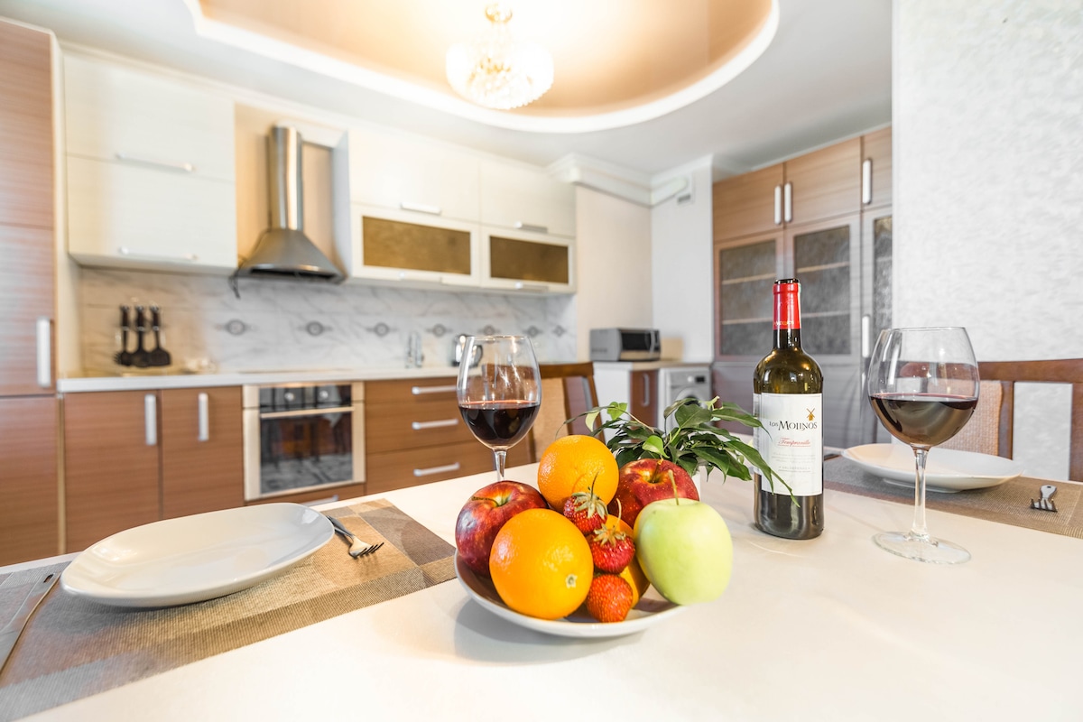 A dining table is set with white plates and wine glasses, accompanied by a bottle of red wine. A bowl filled with fresh fruits, including apples, oranges, strawberries, and a green apple, is positioned at the center. The modern kitchen with wooden cabinets and a stylish exhaust hood is visible in the background.