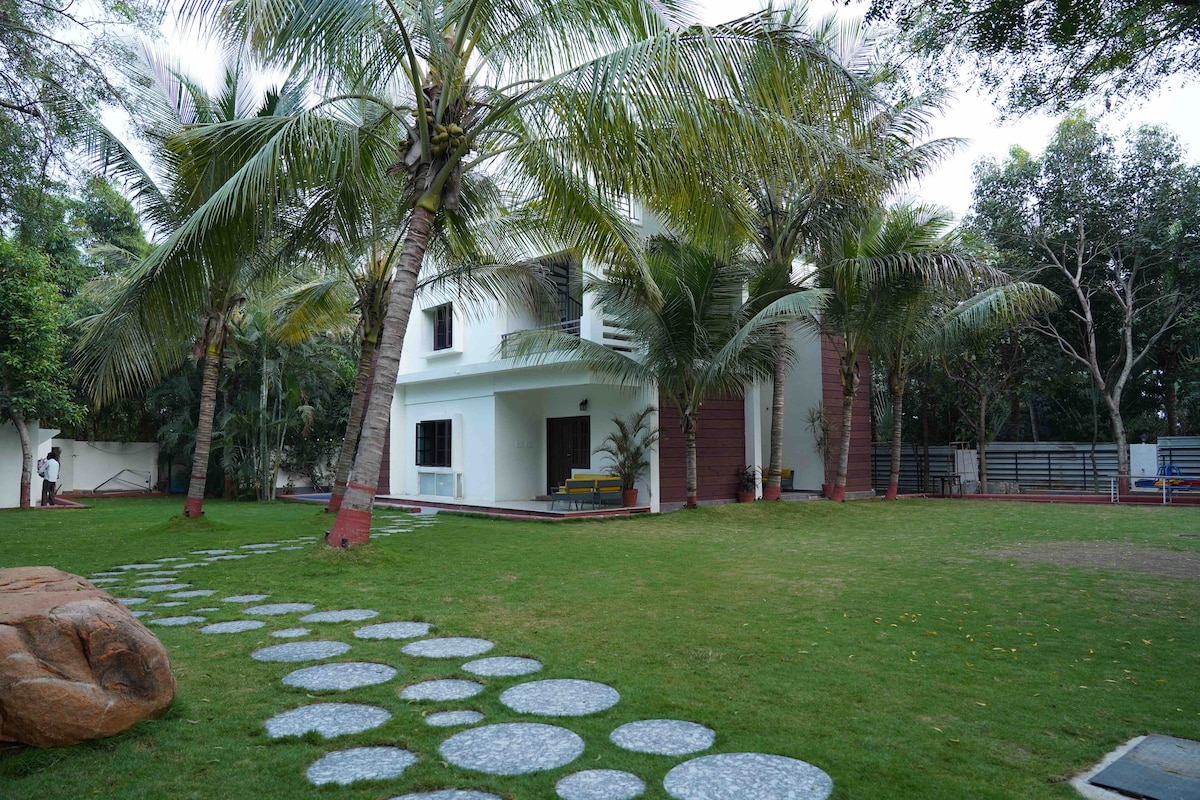 The exterior of a two-story villa is surrounded by lush greenery and tall palm trees. A well-maintained lawn features stone pathway stepping stones leading to the entrance. The building's white facade is complemented by dark accents and large windows, offering a serene and inviting atmosphere.