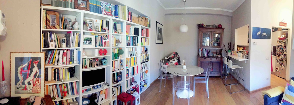 A well-organized living space features a white bookshelf filled with a variety of colorful books and decorative items. A round dining table with transparent chairs occupies the center, while a vintage cabinet adds character to the room. Soft natural light brightens the area.