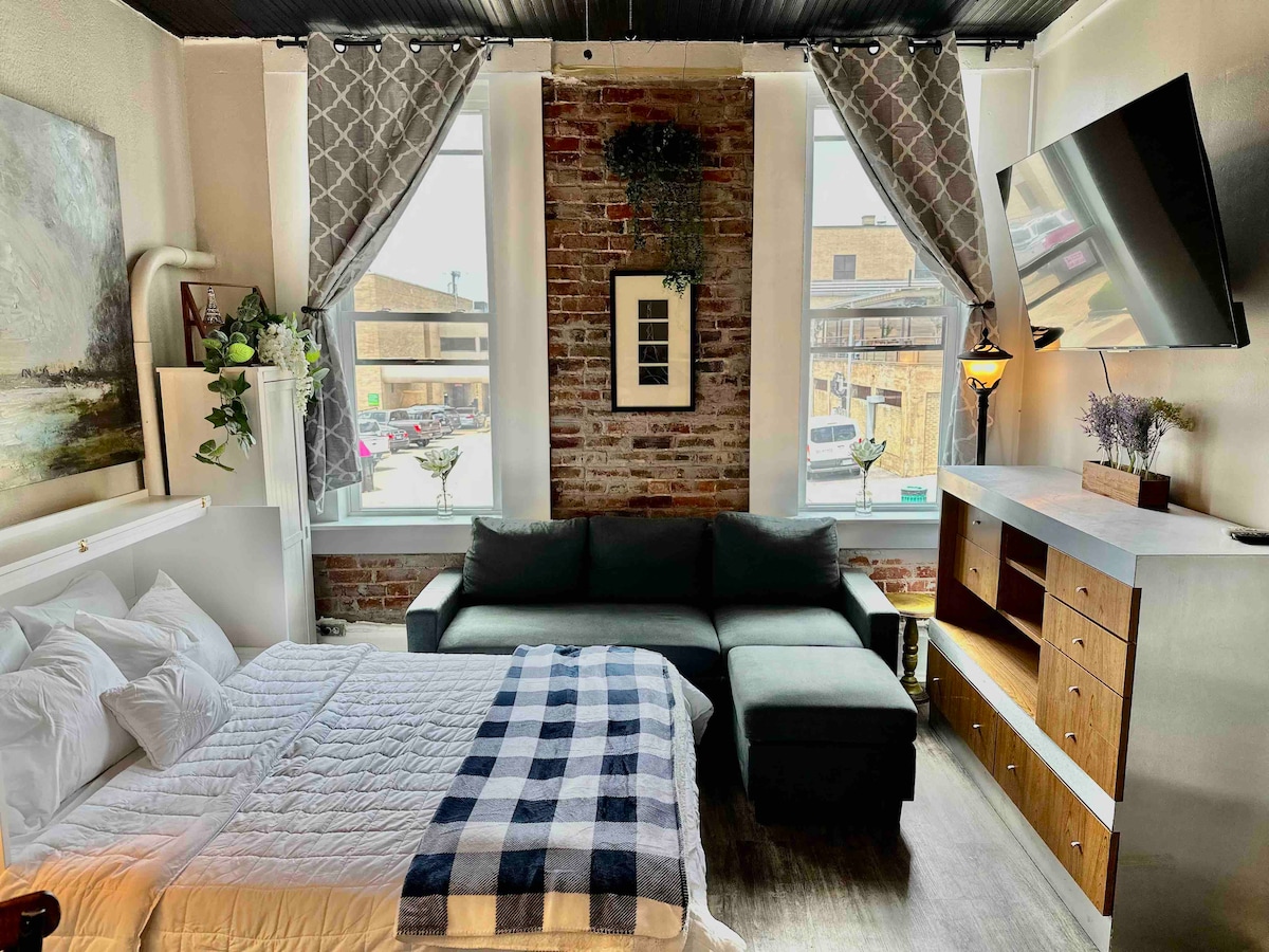 A studio loft features charming brick walls and bright windows that allow natural light to fill the space. A spacious bed is positioned beside a comfortable sectional sofa. A media console and decorative accents enhance the cozy atmosphere, creating a welcoming environment.