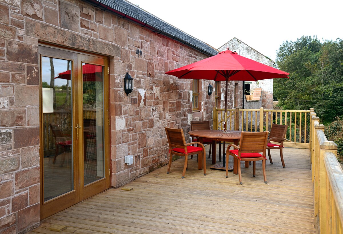 A spacious wooden deck features a round table with seating for six, accompanied by a large red umbrella for shade. French doors lead into the interior, while the stone walls provide a rustic backdrop. Lush greenery is visible in the surrounding area.