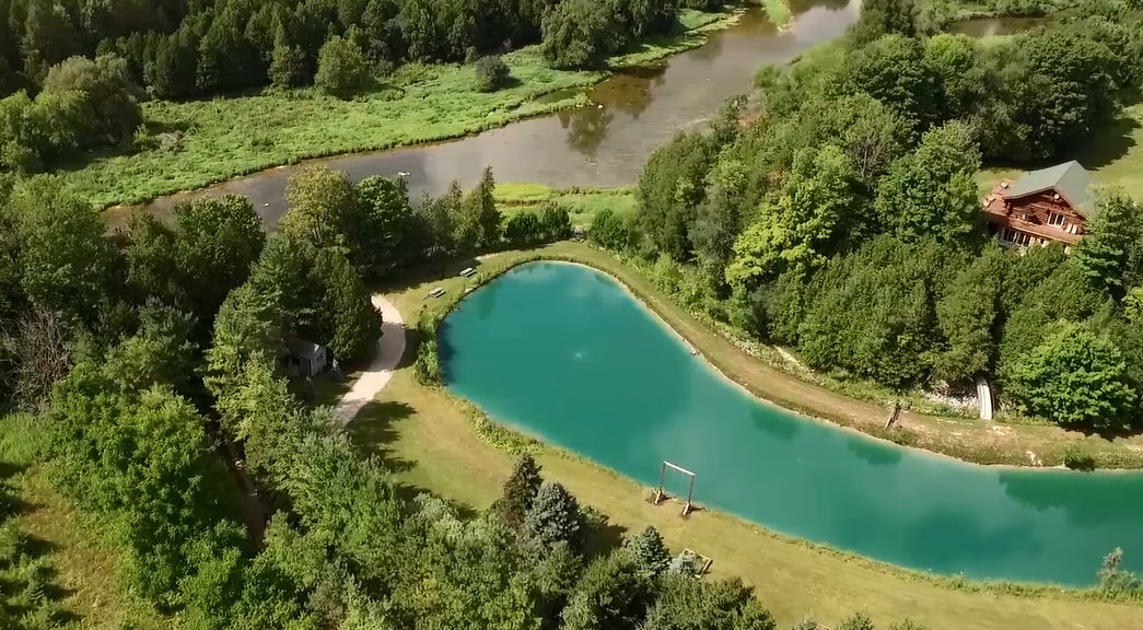An aerial view captures a serene landscape featuring a curving swimming pond surrounded by lush greenery. The tranquil water reflects the surrounding trees, with a small play area visible nearby. The Maitland River meanders gracefully in the distance, enhancing the natural beauty of the property.
