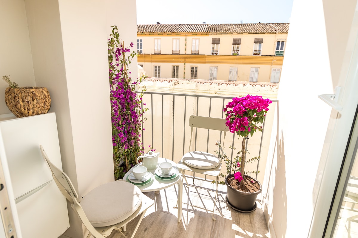 A small balcony area features a round table set for two, accompanied by two chairs. Brightly colored bougainvilleas cascade from pots, adding vibrancy to the space. The balcony provides a view of neighboring buildings with terracotta roofs and balconies adorned with plants.