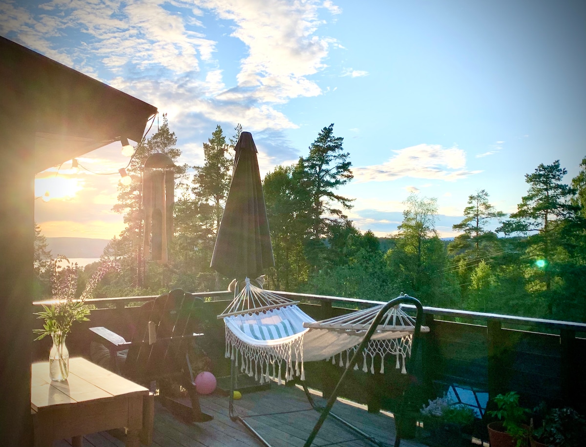 A spacious terrace is shown, illuminated by soft sunlight. A hammock hangs between two supports, with a dark umbrella positioned nearby. Surrounding trees add a natural backdrop, while a wooden table and chairs offer seating options. Scenic views of the fjord and sky are visible in the distance.