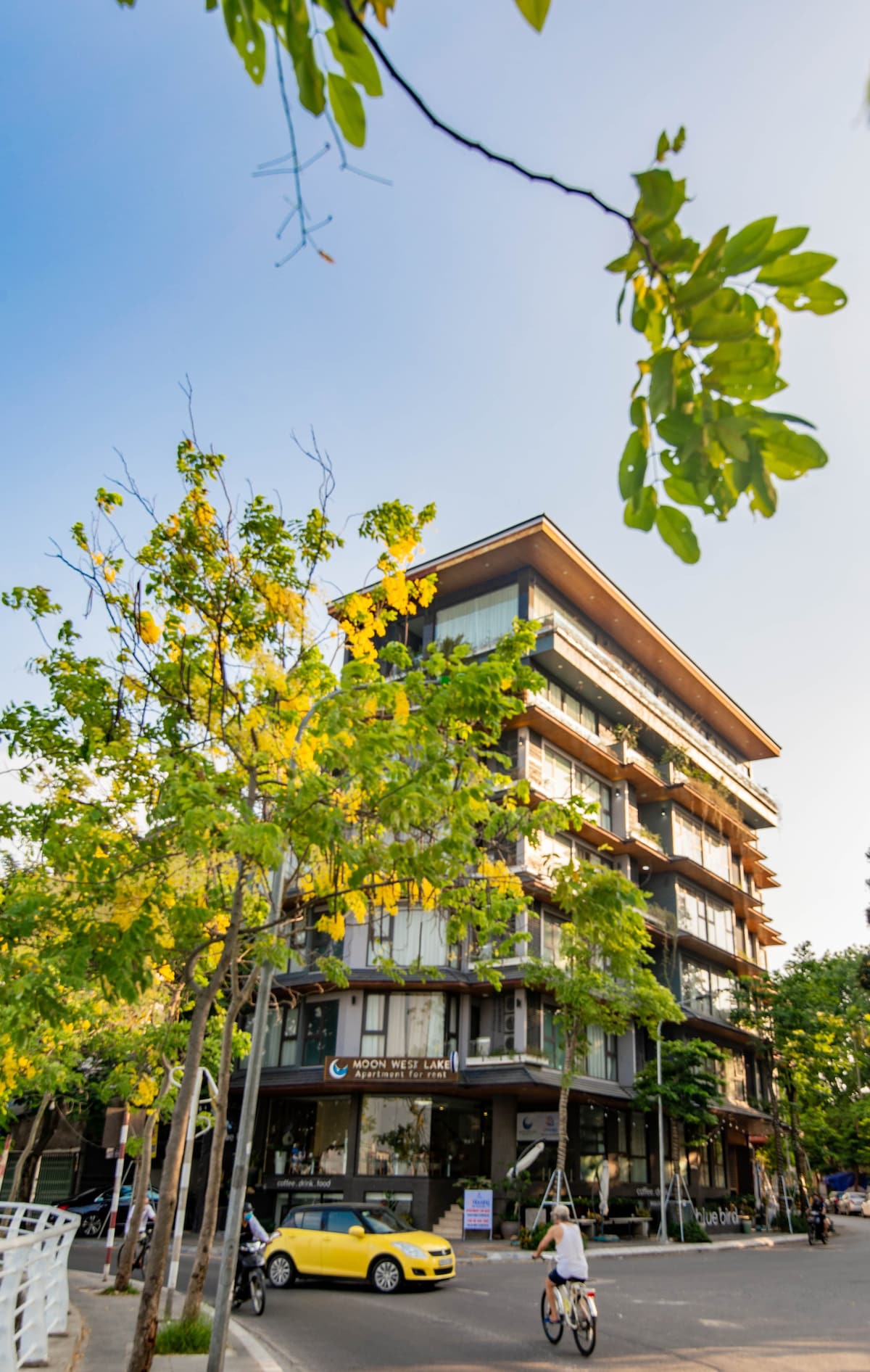 The modern four-story building is surrounded by vibrant greenery and trees. Large windows provide ample natural light, while a welcoming entrance is visible at the ground level. The street includes a parked yellow taxi and a cyclist, enhancing the lively atmosphere of the area.