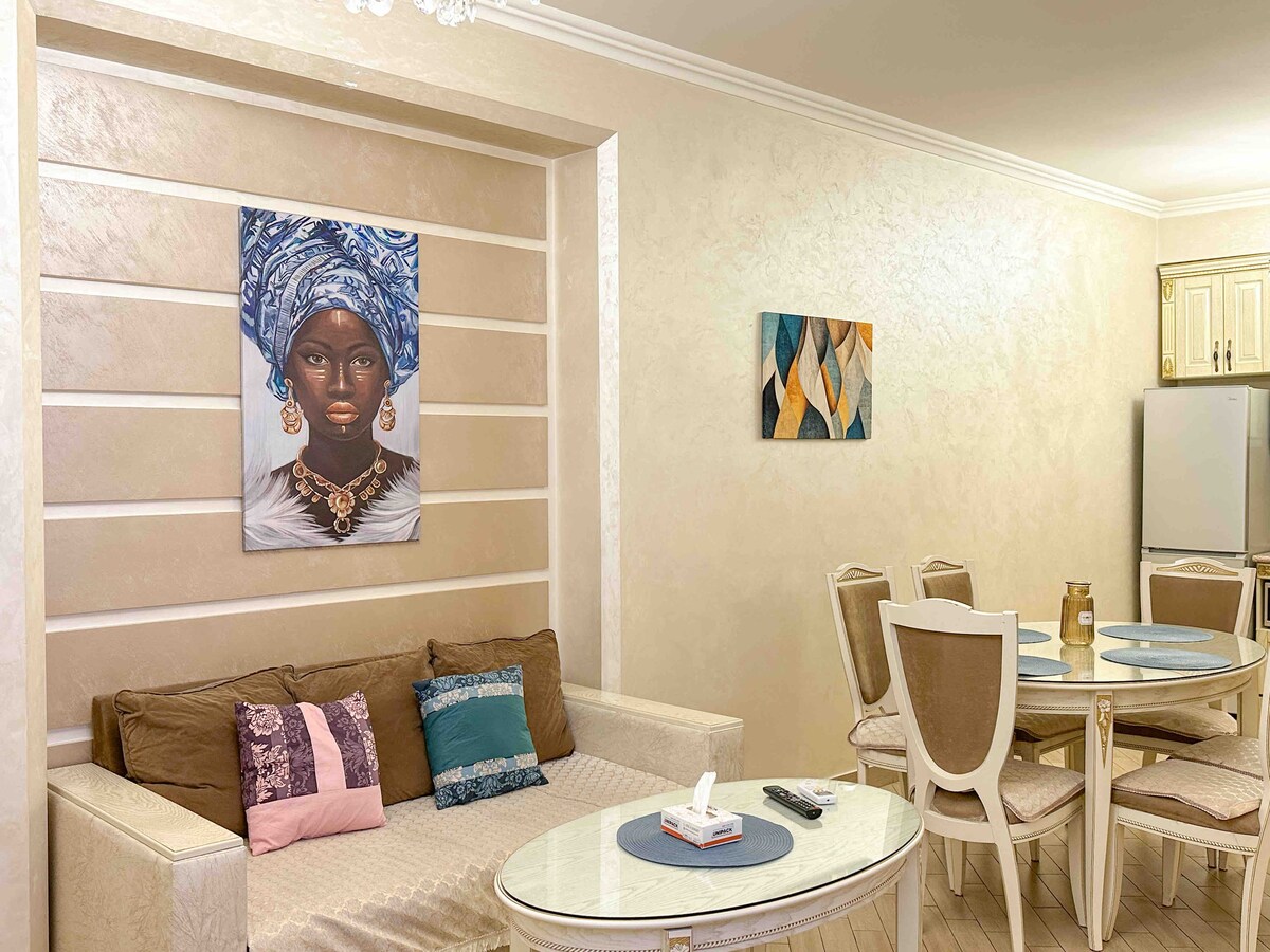 A light-colored living space is highlighted by a comfortable sofa adorned with decorative pillows. A glass coffee table is positioned in front of the sofa. A small dining area features a round table with four elegant chairs. Wall art adds character to the room.