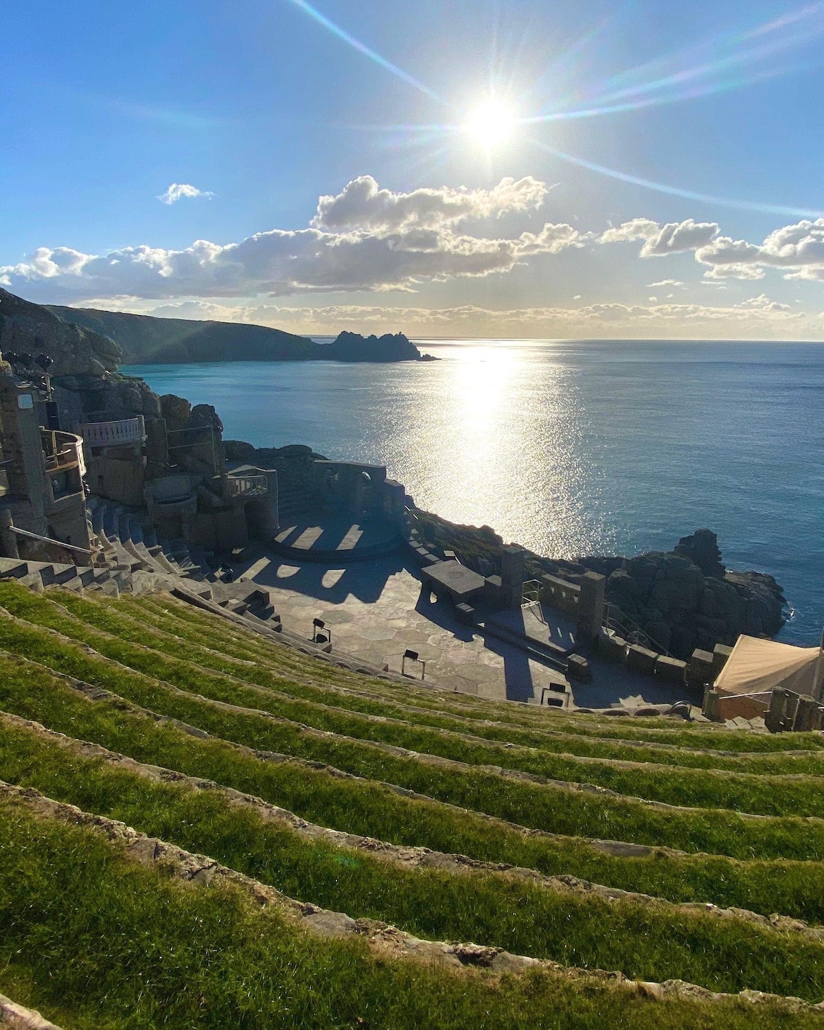 An open-air theater, set against the backdrop of a shimmering sea and dramatic cliffs, features grassy seating areas and stone structures. Sunlight glistens on the water, casting shadows across the landscape. The sky is adorned with soft clouds, enhancing the tranquil atmosphere.