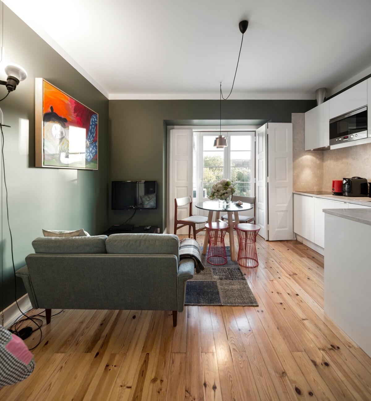 A cozy dining area features a round table set for three, surrounded by simple wooden chairs. A modern green sofa is positioned nearby, complemented by a colorful wall art piece. Natural light filters in through the large open doors, illuminating the wooden flooring.