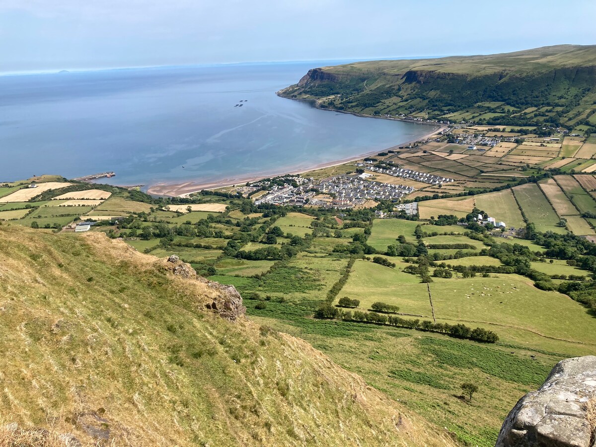 A panoramic view captures the coastline of Waterfoot village, with the tranquil sea and sandy beach stretching along the shore. Green fields and cultivated land create a patchwork effect, while the village is visible in the foreground. The Scottish mainland can be seen in the distance.