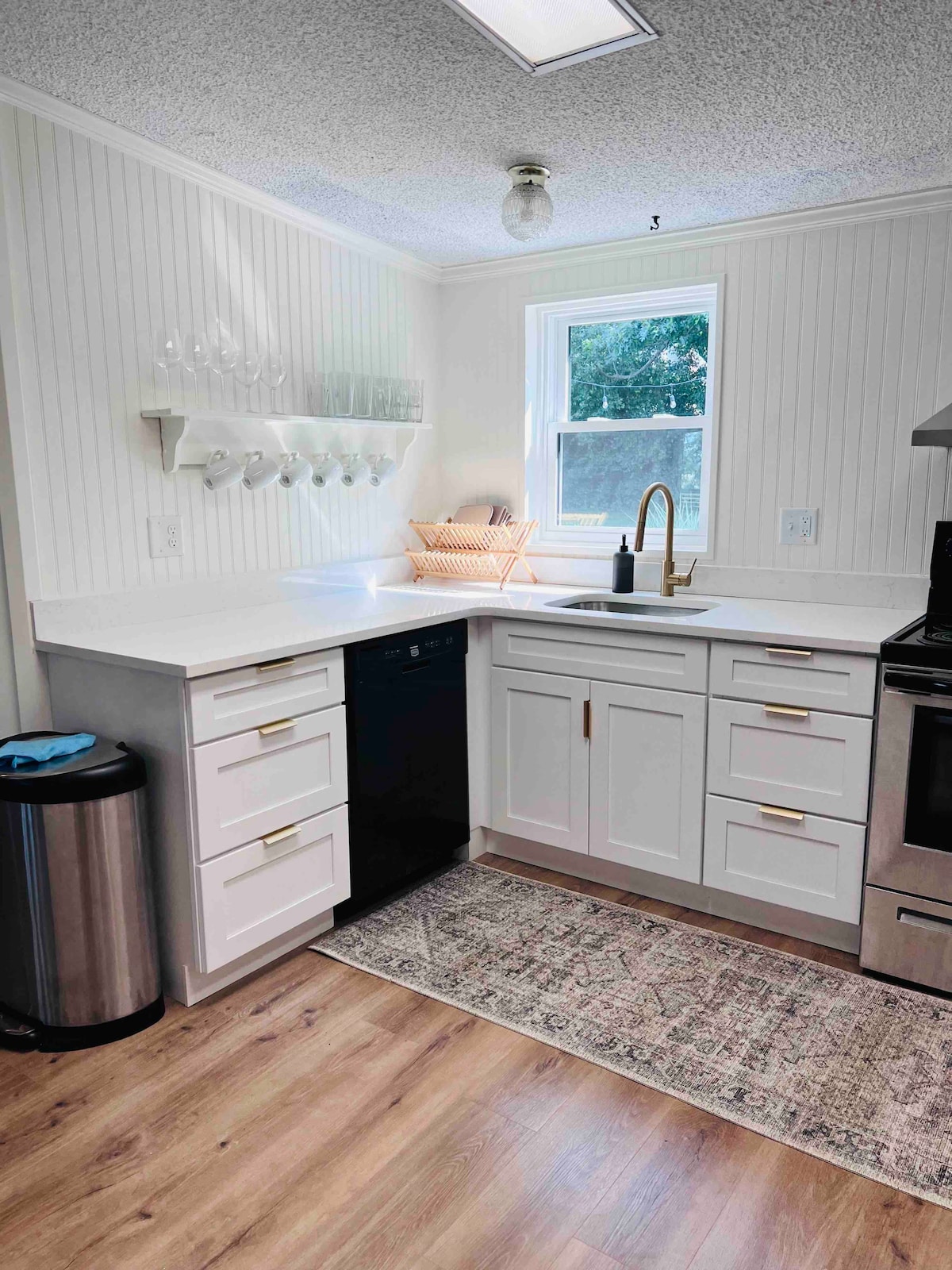 A modern kitchen includes white cabinets with gold hardware, a black dishwasher, and stainless-steel appliances. Glassware is neatly displayed on the wall, and a window provides natural light. A decorative rug lies beneath the sink area, enhancing the functionality of the space.