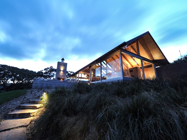 Modern hillside home with large glass windows, illuminated at dusk, showcasing a wooden roof and an outdoor chimney.