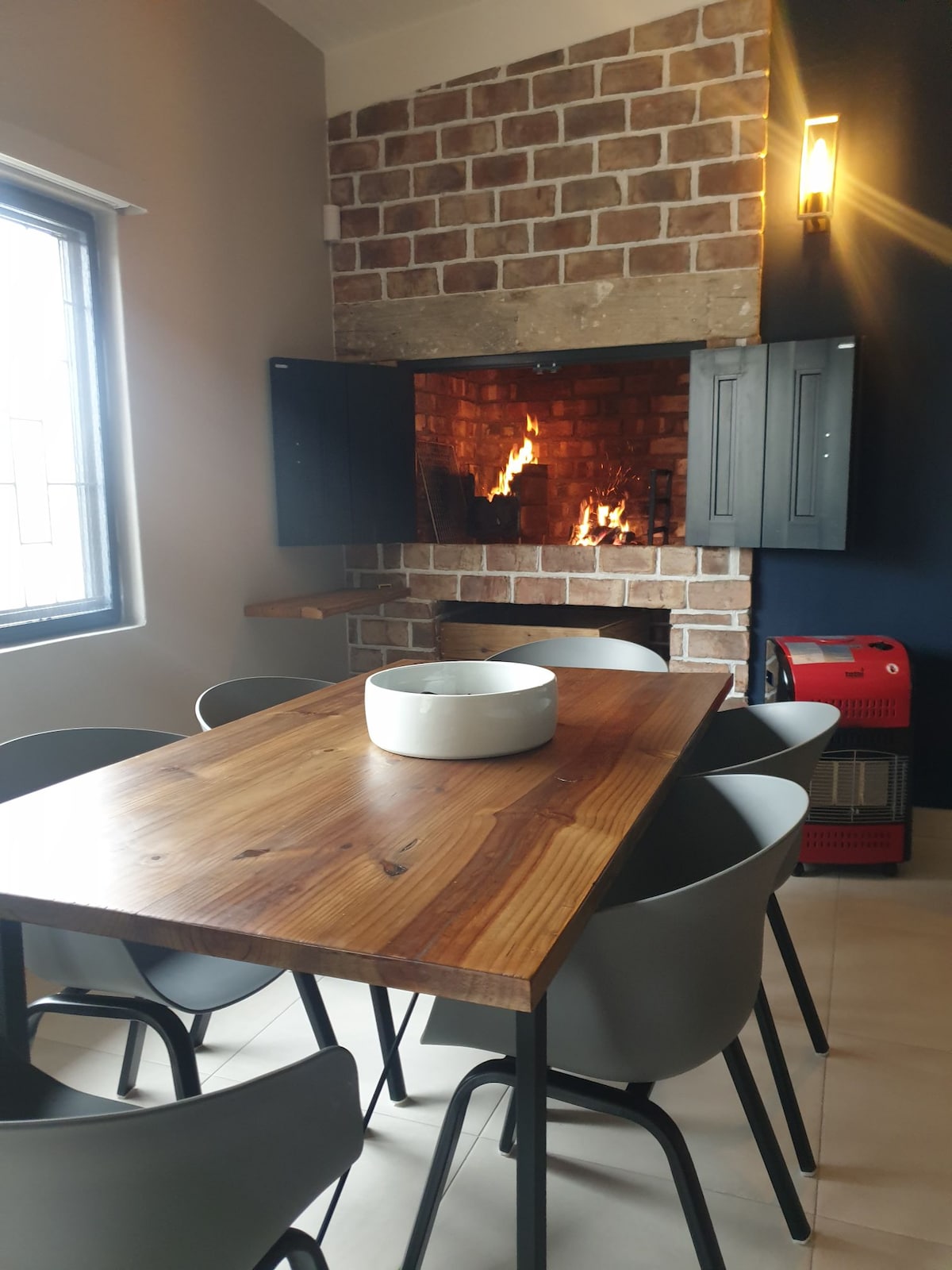 A wooden dining table is surrounded by six grey chairs, creating a communal space. A stone fireplace is visible in the background, featuring a warm fire. Shelving units are positioned beside the fireplace, offering a functional and cozy ambiance.