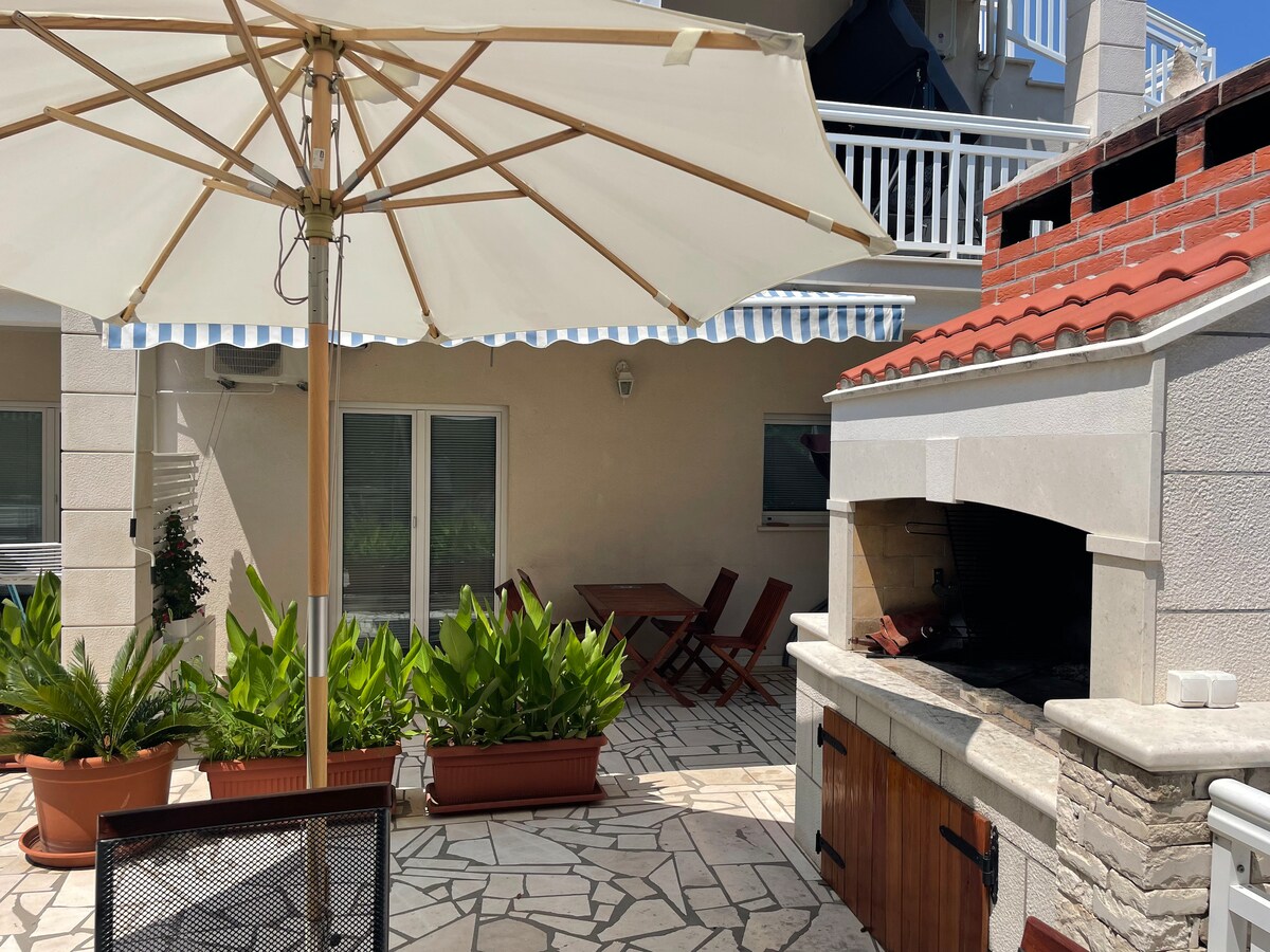 An outdoor terrace features a large umbrella providing shade over a wooden dining table with chairs. Surrounding foliage includes potted plants, and a built-in stone grill is visible on one side, enhancing the available outdoor cooking and dining space.