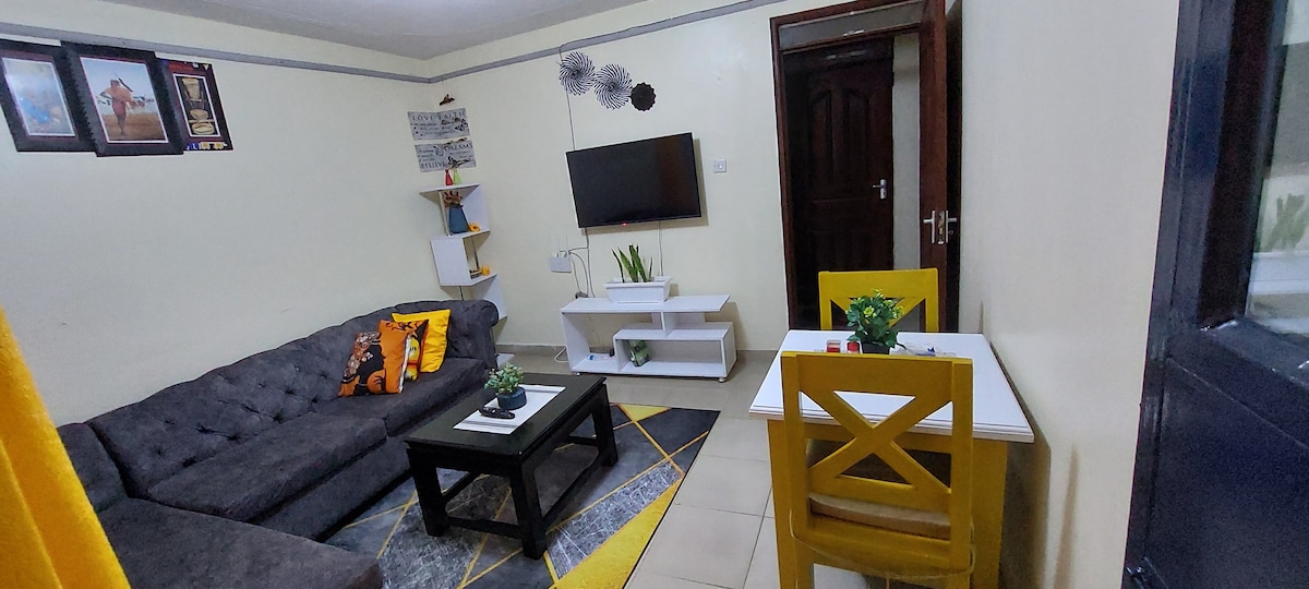A cozy living area features a dark gray sofa adorned with decorative pillows. A TV is mounted on the wall opposite a white table, positioned on a geometric-patterned rug. A yellow chair complements the minimalist decor, along with a few green plants enhancing the atmosphere.
