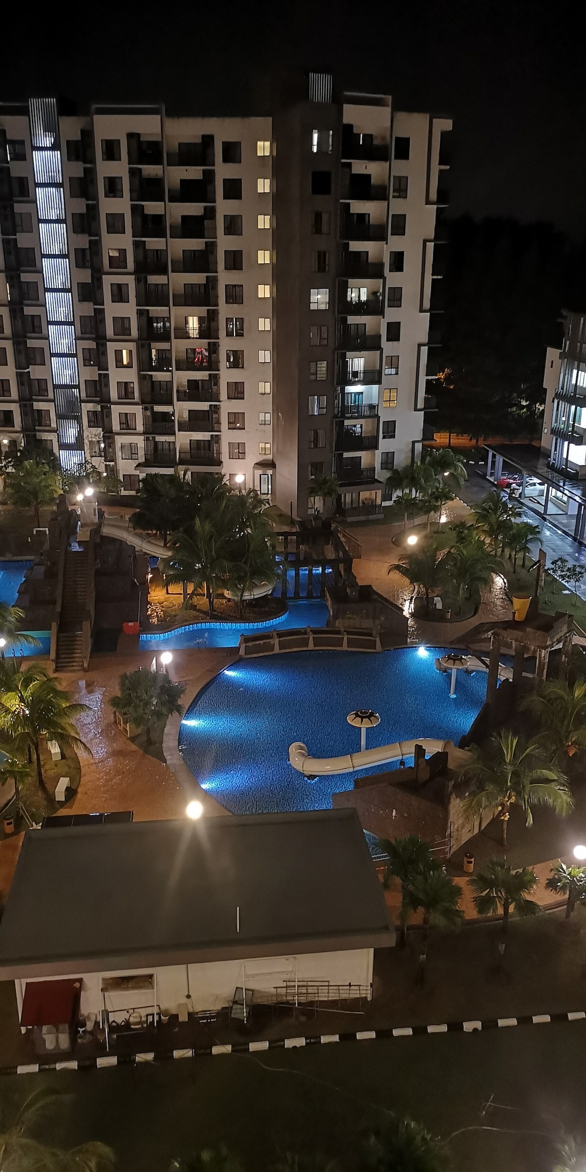 A nighttime view of the property showcases a beautifully illuminated pool area surrounded by lush palm trees. Multiple swimming pools can be seen, including features for children. Soft lighting highlights the architecture of the surrounding buildings, creating a serene and inviting atmosphere.