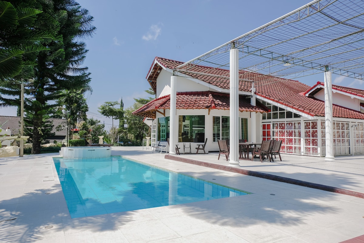 A modern villa is showcased with a clear swimming pool gleaming under the sunlight. Surrounding the pool are lounge chairs and a spacious patio area, complemented by lush greenery and well-maintained trees, enhancing the overall serene environment.