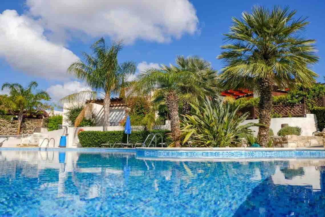 The image showcases a serene communal pool surrounded by lush palm trees and well-maintained gardens. The water reflects the clear blue sky, creating a tranquil ambiance. Lounge chairs are positioned nearby, offering a place for relaxation under the sun.