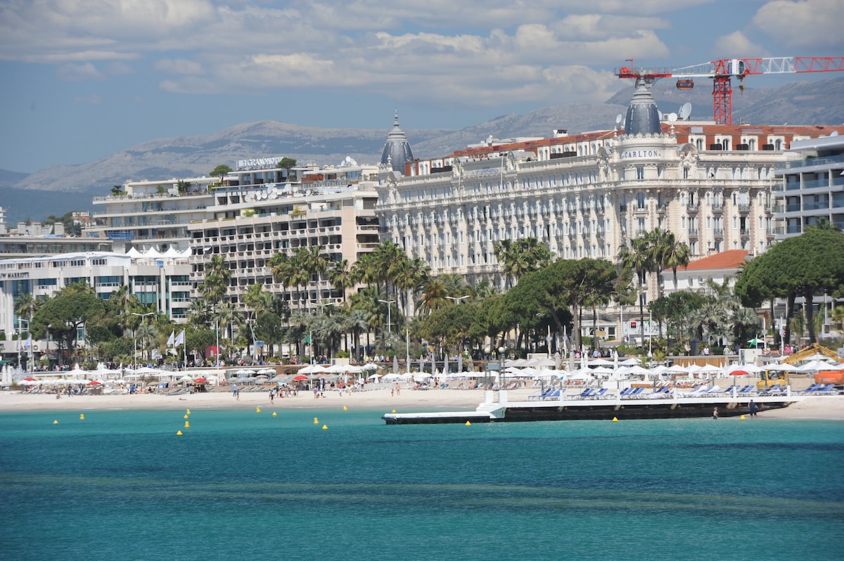 A view of the coastline features a sandy beach populated with sun loungers and umbrellas. The clear turquoise water contrasts with the architecture of surrounding hotels, highlighted by palm trees. The background captures a distant line of mountains under a sky dotted with clouds.