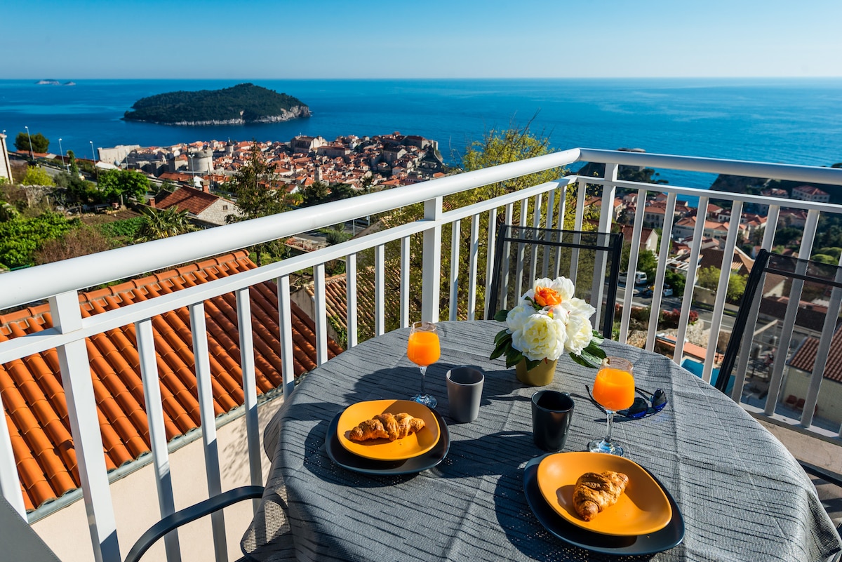 A well-set table is positioned on a balcony, featuring two plates with croissants, colorful drinks, and a floral centerpiece. In the background, a panoramic view showcases the Adriatic Sea and the coastline, with the charming rooftops of Dubrovnik visible.