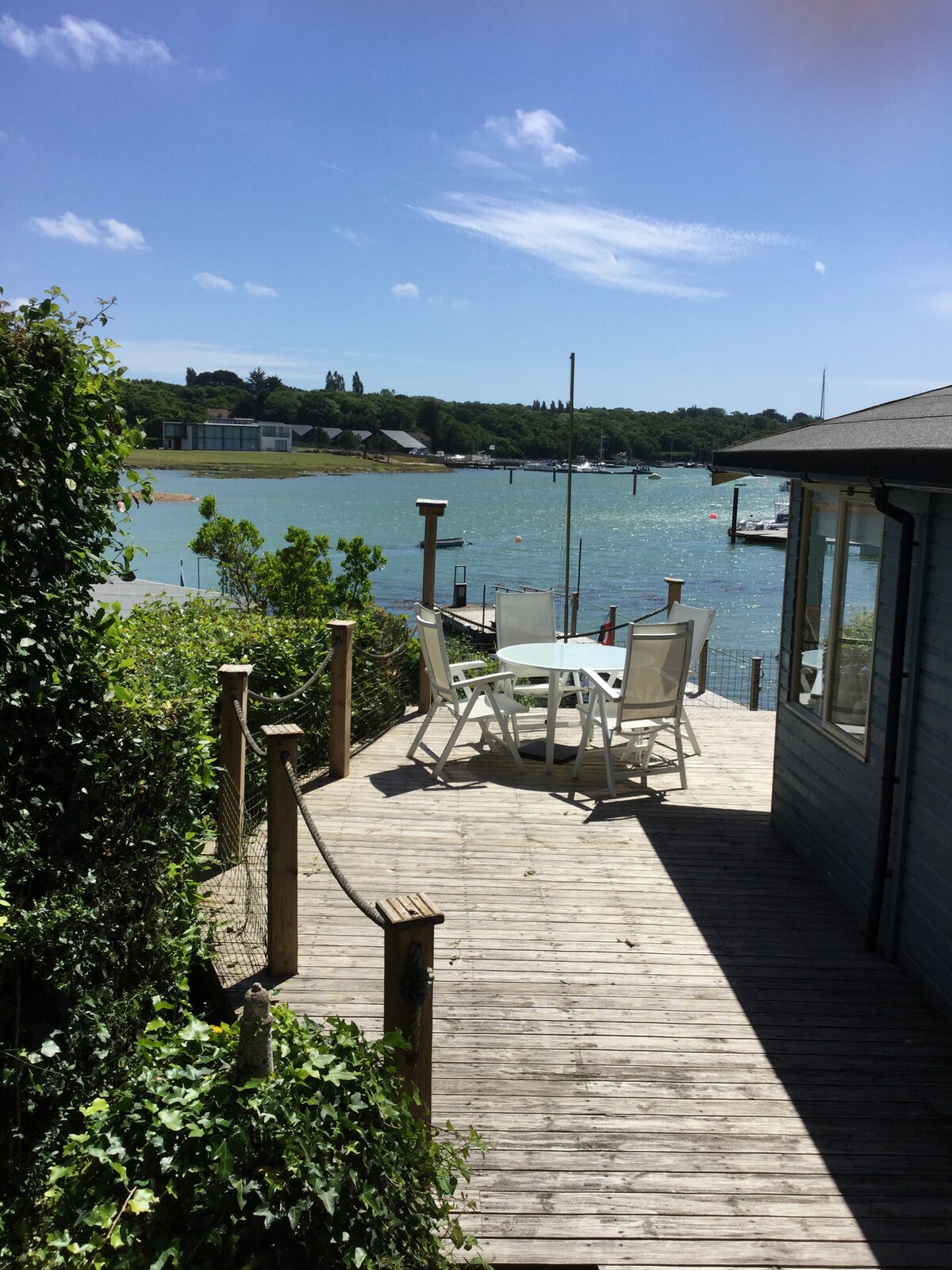 A wooden deck is visible, equipped with a round table and four white chairs overlooking the water. Lush greenery frames the space, while the bay's blue waters and distant boats can be seen under a clear sky.