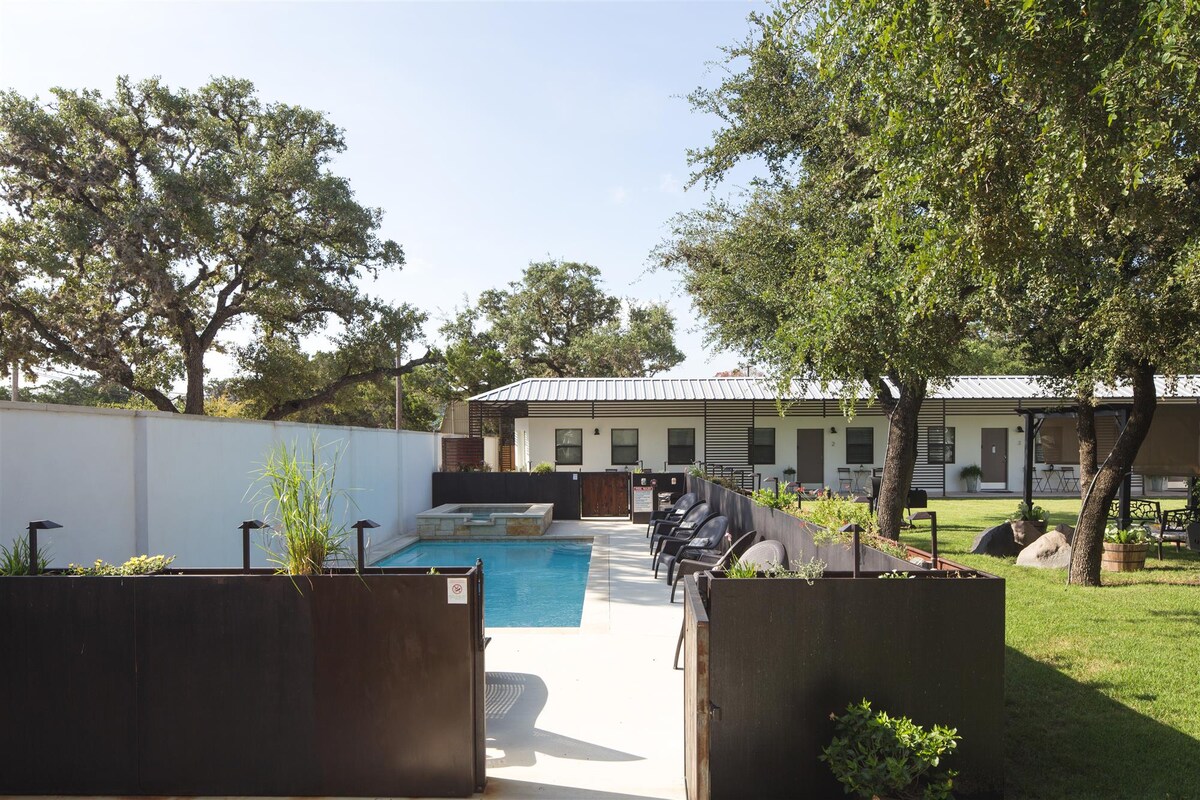 A communal outdoor area features a well-maintained pool surrounded by lounge chairs and greenery. The setting is enclosed by a low wall, with an accessible path leading to a building featuring several entries. Shade is provided by mature trees nearby.