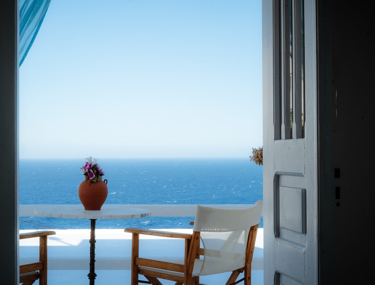A serene balcony is featured, with a small round table and two wooden chairs positioned to face the expansive Aegean Sea. A terracotta pot with pink flowers adds a subtle touch of color. Bright blue skies merge with the ocean in the background.