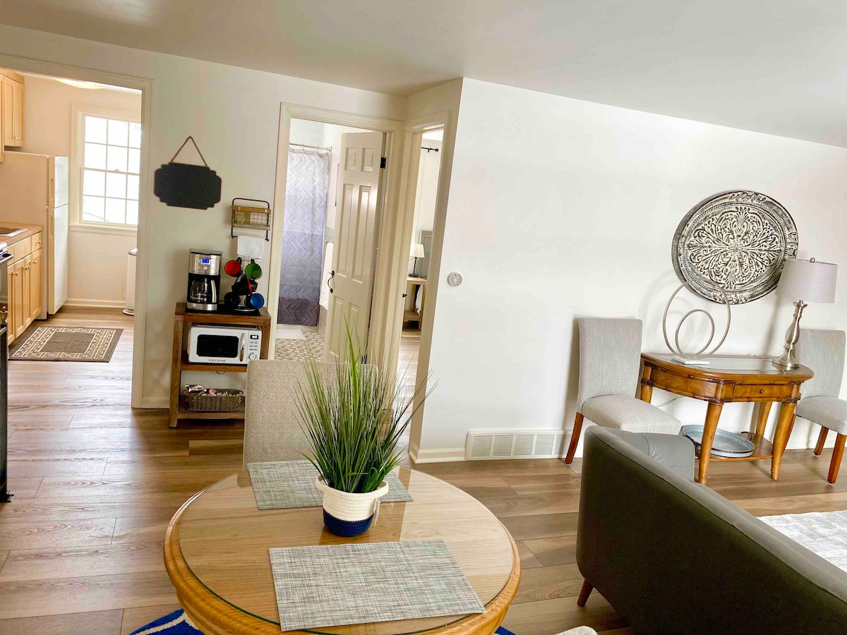 An open living space features a round wooden dining table set for two, accompanied by a decorative plant. A gray couch is positioned to one side, with soft light illuminating the area. The room connects to the kitchen and a bathroom area is visible in the background.