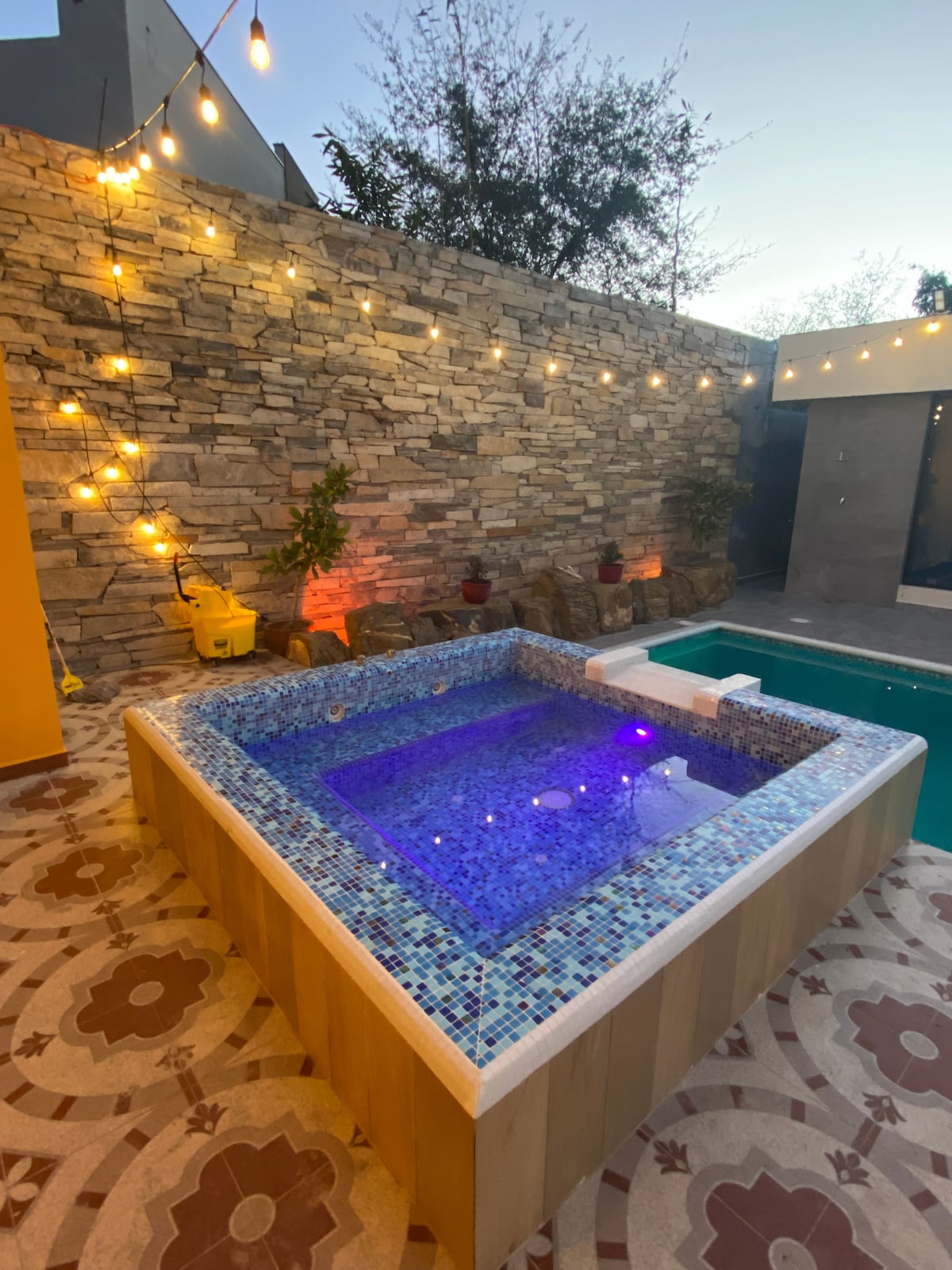 A jacuzzi is situated outdoors, surrounded by a decorative stone wall and string lights that create a cozy ambiance. The mosaic tiles reflect water in soft hues, complementing the nearby pool. A decorative floor pattern enhances the inviting outdoor space.