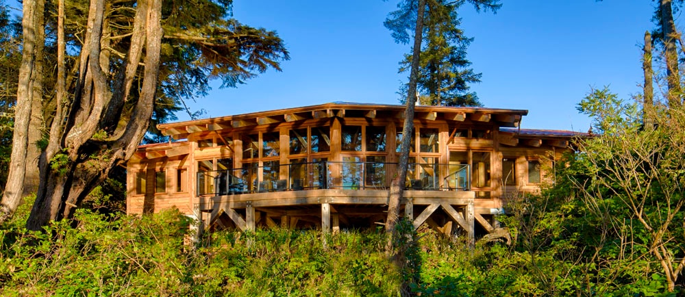 This cedar-built lodge is set amongst lush greenery, elevated on cement stilts. Expansive windows bring in natural light and offer views of the surrounding forest. A spacious deck is visible, framed by trees, ideal for enjoying the outdoor environment.