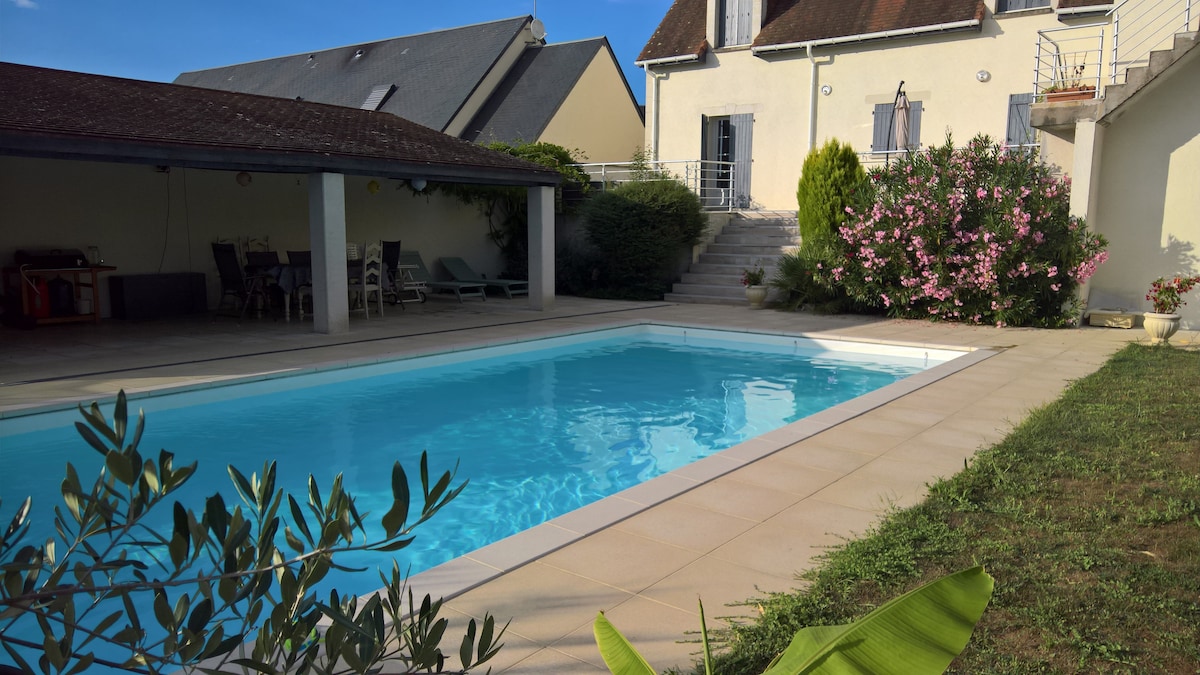 A serene outdoor space features a swimming pool surrounded by a tiled patio. Lush greenery is present along the edges, with flowering shrubs adding color. A shaded area with seating can be seen in the background, and a two-story house provides structural interest against a clear blue sky.