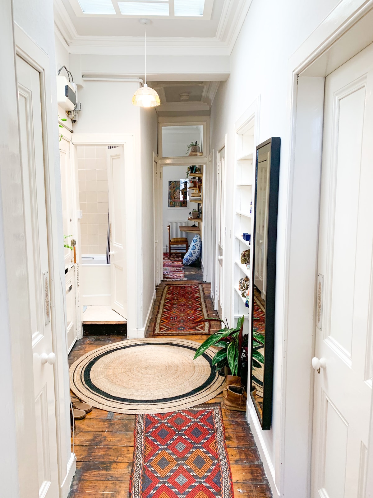 A well-lit hallway is lined with white doors and features vibrant red rugs. Natural light enters through a skylight, enhancing the wood flooring. Decorative plants and shelves add character, while a circular mat centers the space, leading towards the inviting rooms.