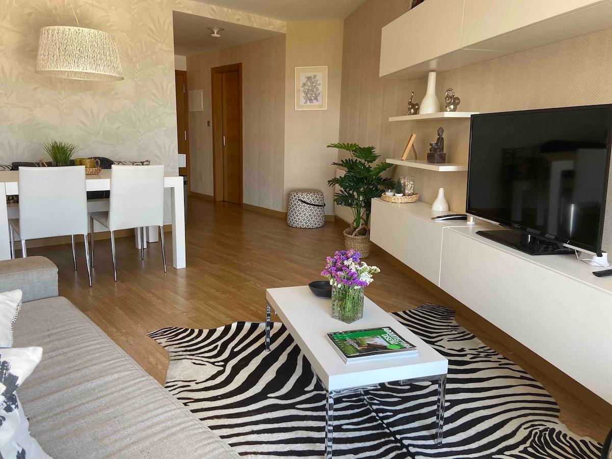 A modern living area features a comfortable sofa in muted tones, accompanied by a glass coffee table atop a zebra-striped rug. A sleek television is mounted on the wall, and a dining table with chairs is visible in the background, complemented by decorative plants.