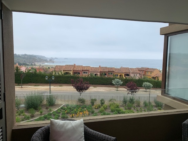 A serene view of the ocean is displayed from the terrace, framed by a glass window. Green plants and colorful flowers line the foreground, while residential buildings with terracotta roofs are visible in the distance under a soft, overcast sky.