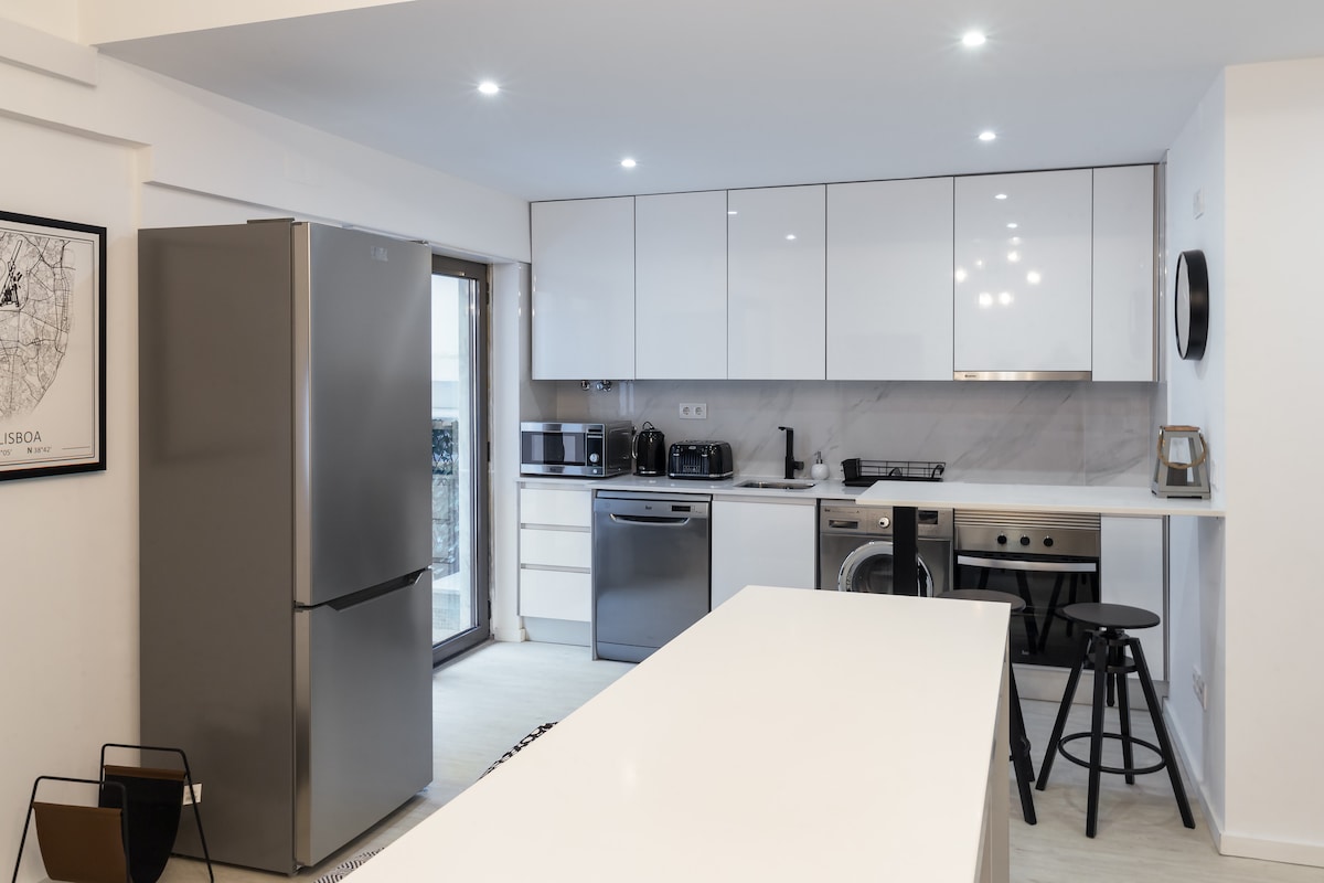 An open-concept kitchen area is showcased, featuring modern cabinetry and sleek appliances. A large silver refrigerator stands alongside a countertop where a bar stool is positioned. Large glass doors provide natural light and access to an outdoor space.