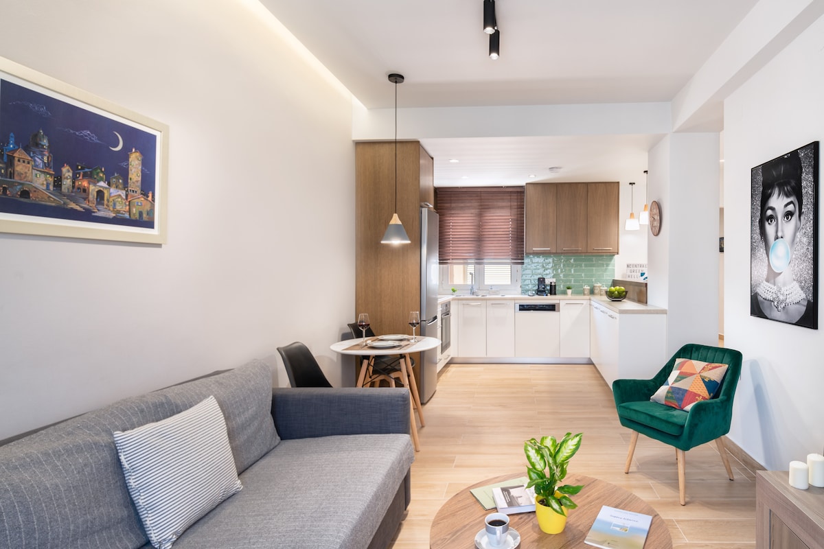 The living area is characterized by a gray sofa facing a wooden coffee table, with a green armchair nearby. A circular dining table with two chairs is positioned against the wall, while the adjacent kitchen features modern cabinetry and a vibrant backsplash. Natural light enters through the windows.