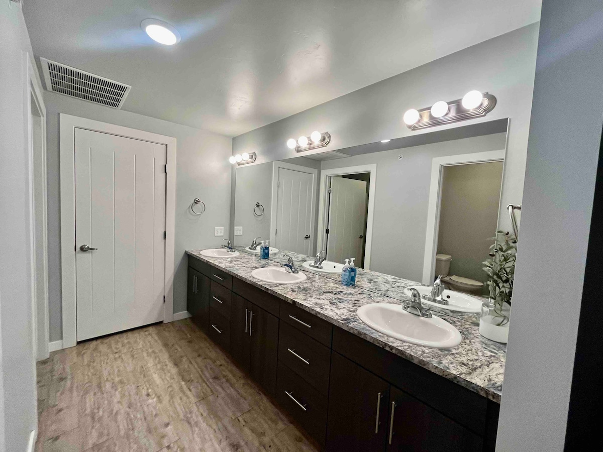 A well-lit bathroom features a long granite countertop with dual sinks and modern fixtures. Stylish mirrors above each sink reflect the space. Soft-colored walls complement the warm tones of the flooring. A separate room is visible in the background, enhancing privacy.