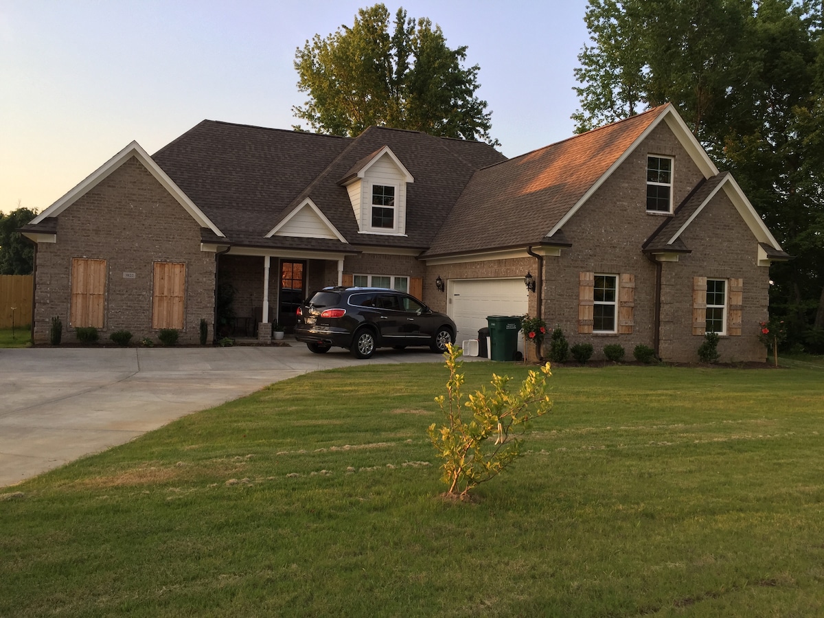 The exterior of a spacious home is depicted, surrounded by a well-manicured lawn. A paved driveway leads to a two-car garage, with greenery lining the path. The house features a blend of brick and siding, accented by large windows and inviting landscaping.