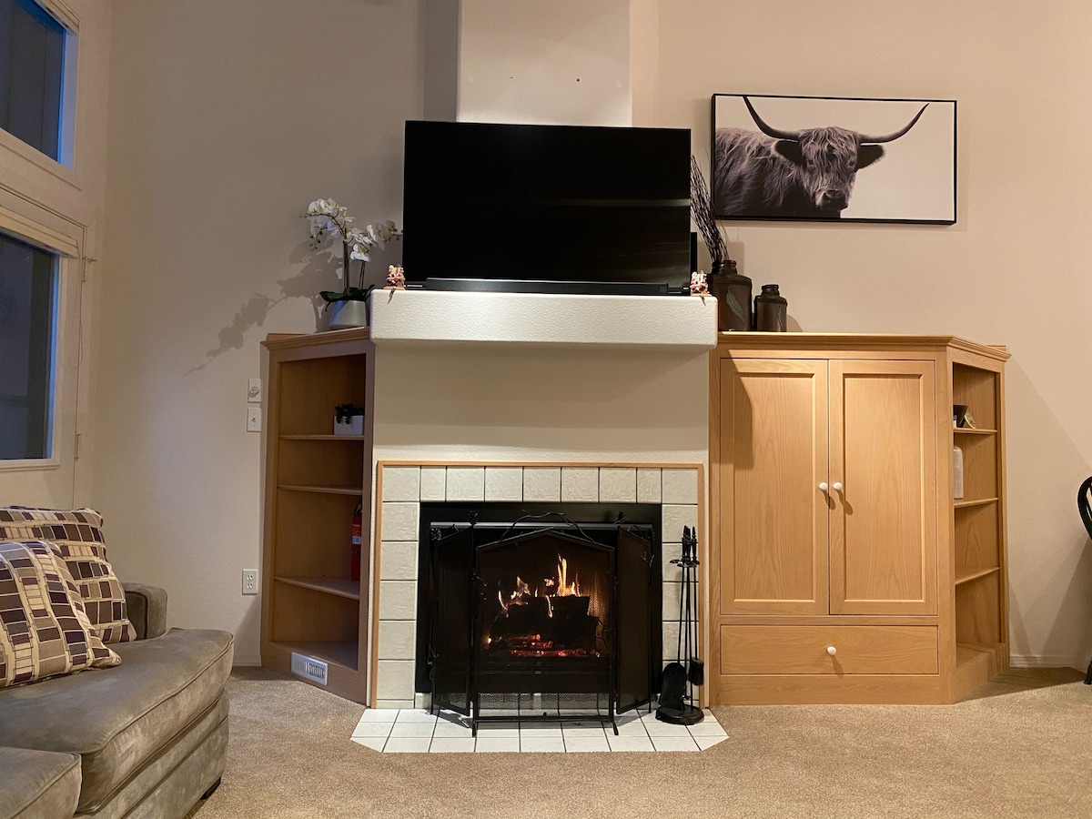 A cozy living area is presented, featuring a stone fireplace with a lit fire. A large flat-screen TV rests above the mantel. Light wood cabinetry and shelves are noticeable, while a comfortable couch, adorned with patterned pillows, offers a welcoming spot to relax.