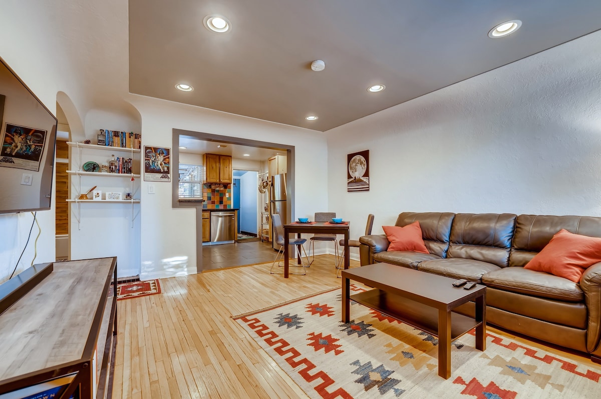 The open living area is well-lit, featuring a comfortable brown leather sofa with orange accent pillows. A wooden coffee table is centered on a patterned rug. The kitchen is visible in the background, showcasing wooden cabinetry and modern appliances.