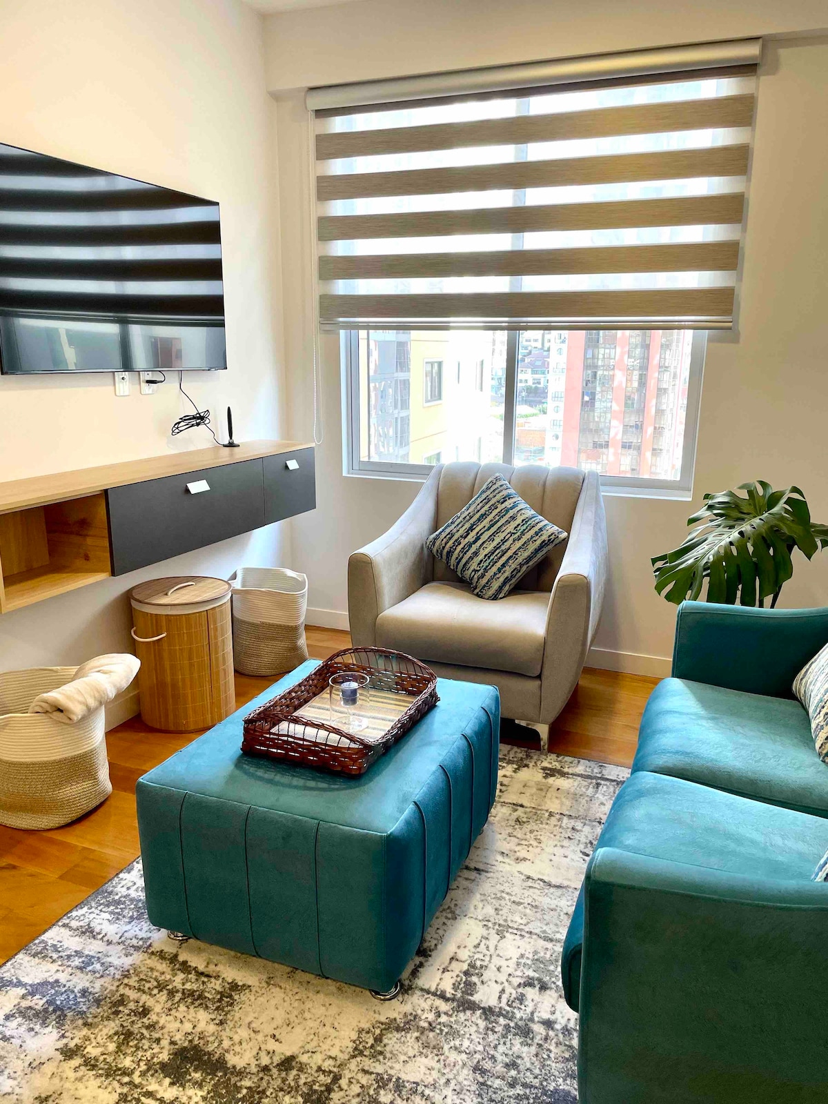The living area features a comfortable turquoise sofa and a light gray armchair. A small coffee table sits at the center, adorned with a tray and decorative items. Natural light filters through striped window shades, enhancing the warm ambiance of the space.