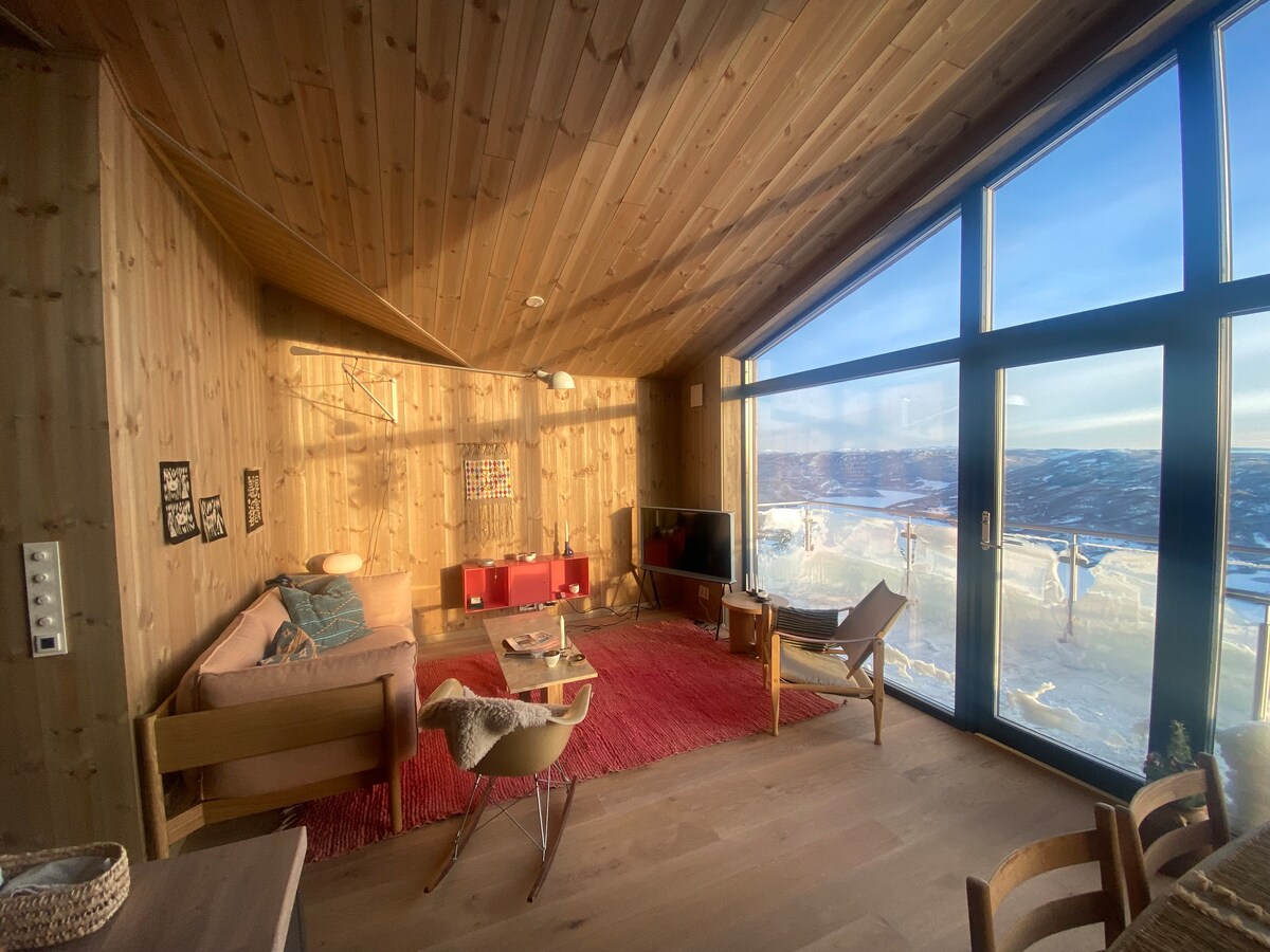 An open living area is highlighted by wooden walls and a sloped ceiling, featuring large windows that offer expansive views of the surrounding landscape. A cozy seating arrangement includes a light-colored sofa and chairs, complemented by a warm area rug. A television is positioned nearby.