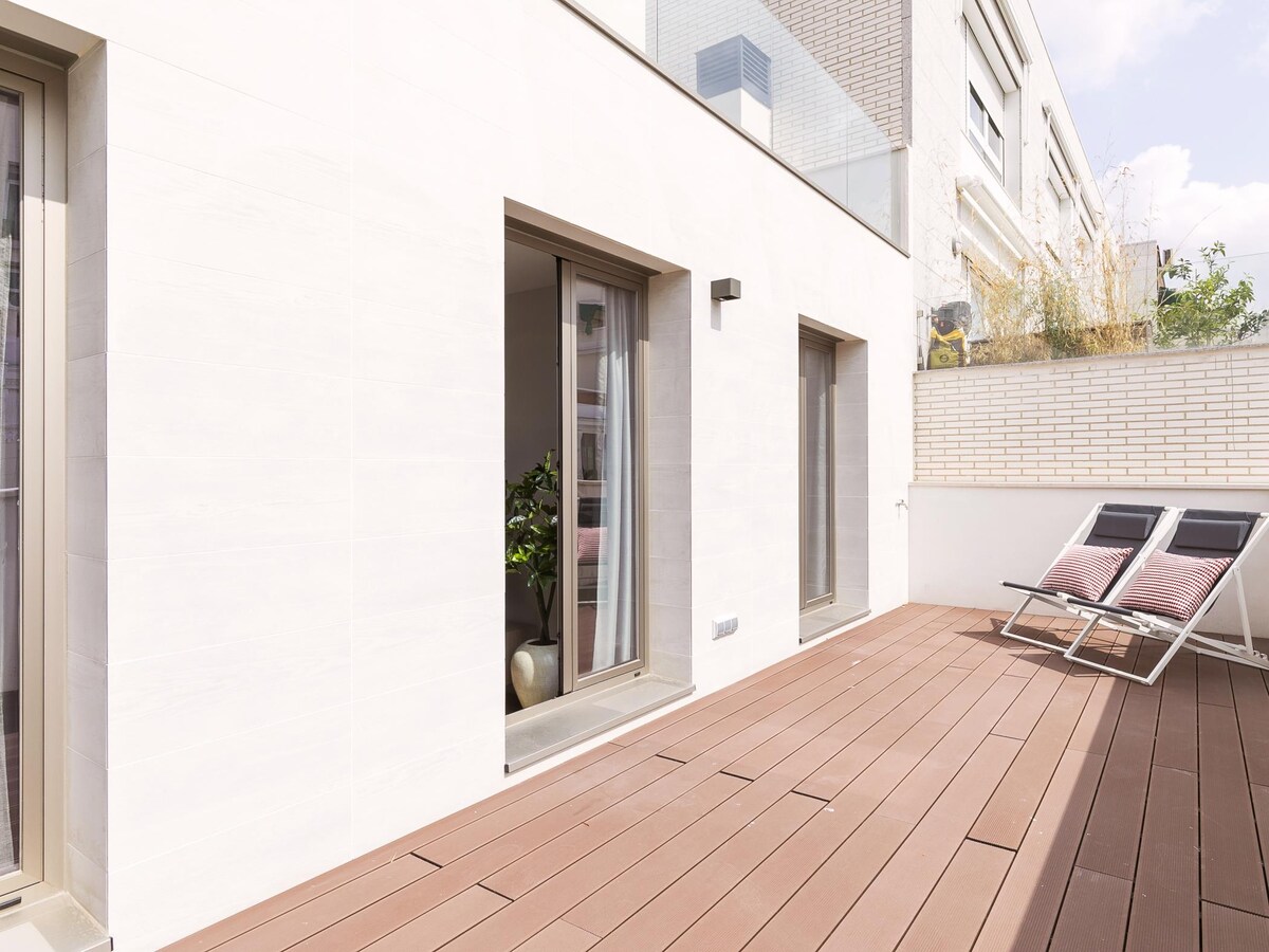 A private terrace features smooth wooden decking, with two loungers positioned for relaxation. Sliding glass doors provide access to the interior space. A minimalist aesthetic is complemented by planters and greenery outside the walls.