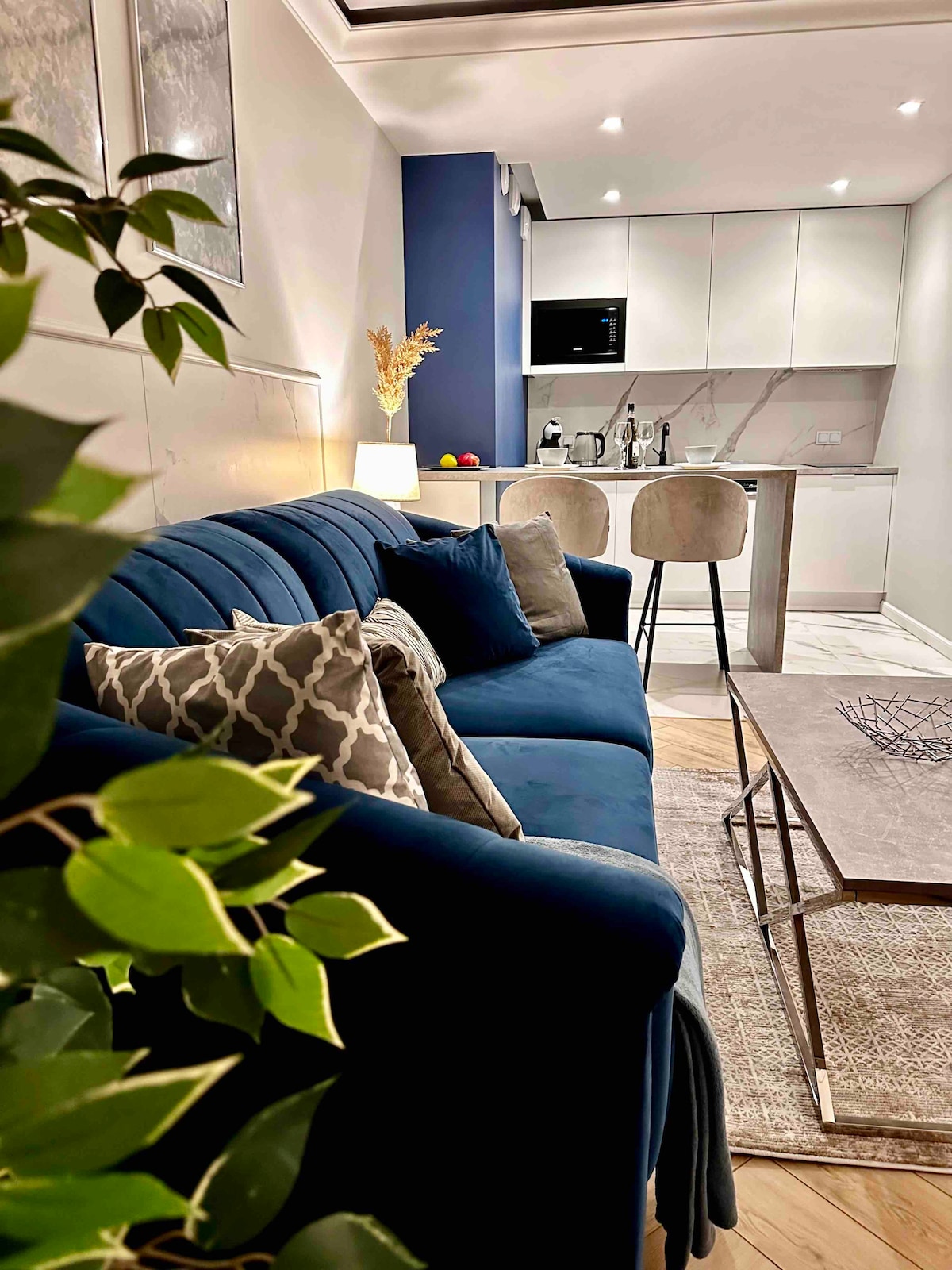A modern living area featuring a deep blue sofa adorned with decorative pillows. A sleek countertop with bar stools is visible, leading to a well-equipped kitchen area. Soft lighting illuminates the space, complemented by greenery nearby.