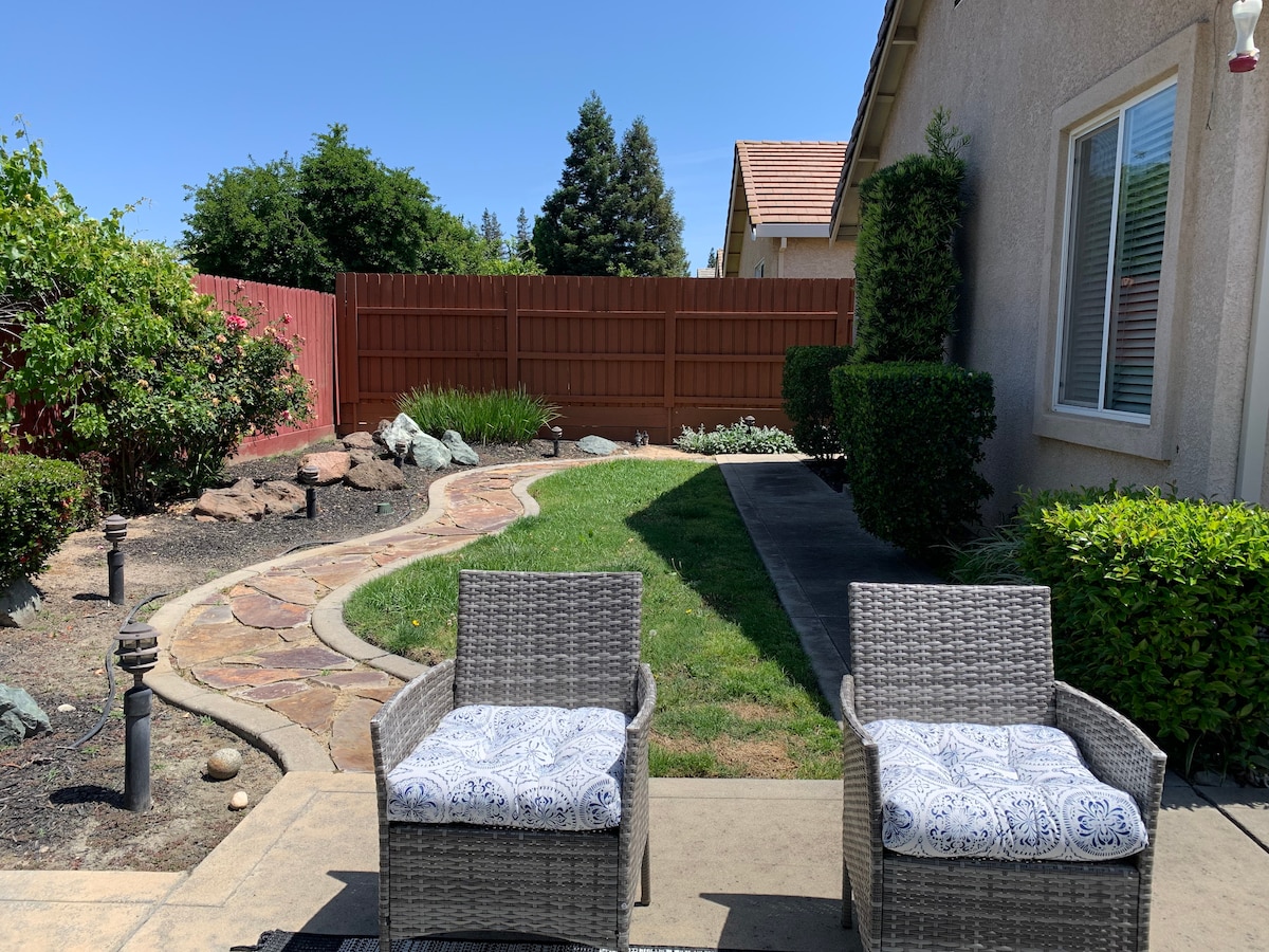 An outdoor patio area features two gray wicker chairs with patterned cushions, positioned on a stone-paved surface. Lush grass and shrubbery are visible, along with a winding pathway made of stone. A wooden fence provides privacy and frames the serene garden space.
