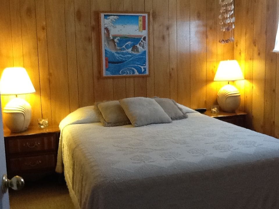 A cozy bedroom features warm wooden paneling complemented by soft lighting from two bedside lamps. A spacious bed is dressed in a light quilt, with two decorative pillows placed atop. A piece of artwork depicting waves is hung above the bed, enhancing the beach theme.